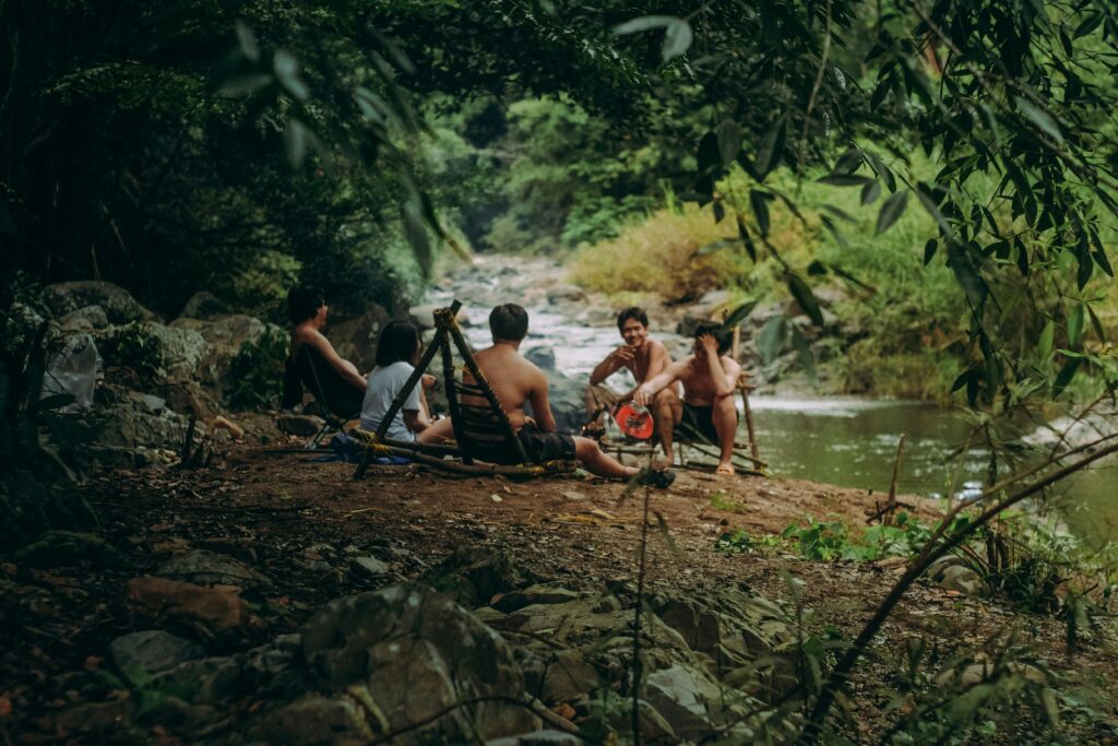 pexels-photo-2888108-2888108 A group of adults relaxing and eating by a riverbank surrounded by lush greenery.